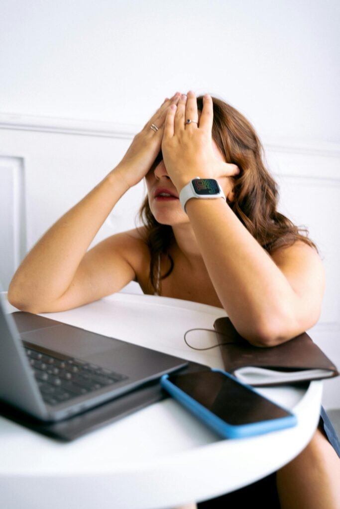 A woman sits at a white round table with her hands over her face, appearing stressed or tired. A black laptop, blue phone, and brown notebook are on the table in front of her.