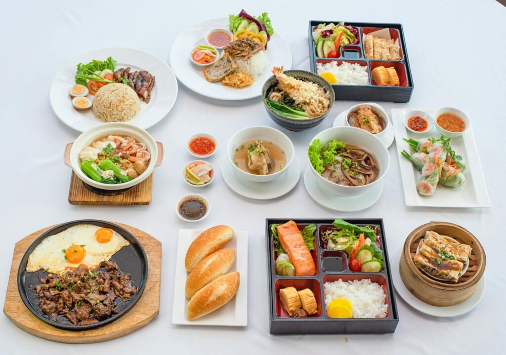A diverse array of Asian cuisine including several rice bowls with various toppings, steaming noodle soup bowls, compartmentalized bento boxes with colorful ingredients, crispy spring rolls, and a sizzling plate featuring beef and eggs, all neatly arranged to showcase the variety of traditional and modern Asian dishes available at the restaurant.