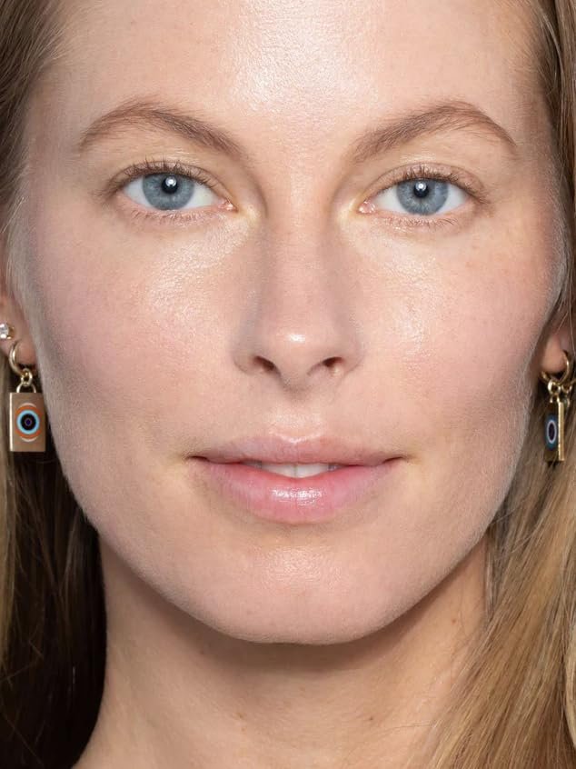 A bottle of ILIA Super Serum Skin Tint SPF 40 in the shade "Bom Bom" lying on its side next to a few pumps of the liquid tint on a white surface, highlighting its lightweight texture.

