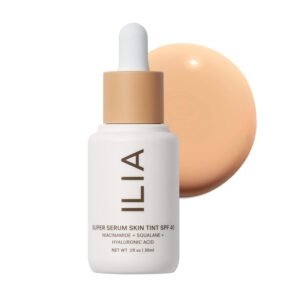 A bottle of ILIA Super Serum Skin Tint SPF 40 in the shade "Bom Bom" lying on its side next to a few pumps of the liquid tint on a white surface, highlighting its lightweight texture.