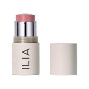 An ILIA Multi-Stick in the shade "Tenderly" swatched on a forearm, showing a soft, creamy pink color, with the stick itself resting nearby on a natural linen surface.
