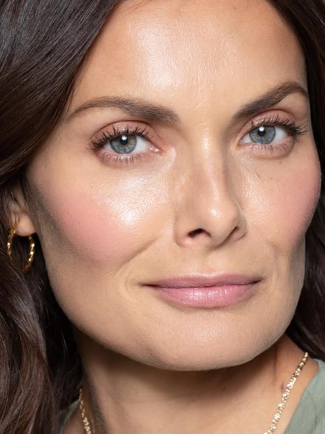 An ILIA Multi-Stick in the shade "Tenderly" swatched on a forearm, showing a soft, creamy pink color, with the stick itself resting nearby on a natural linen surface.