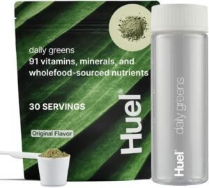 A glass of vibrant green Huel Daily Greens is next to the original bag and a clear Huel bottle, with fresh apples and mint leaves in the background.