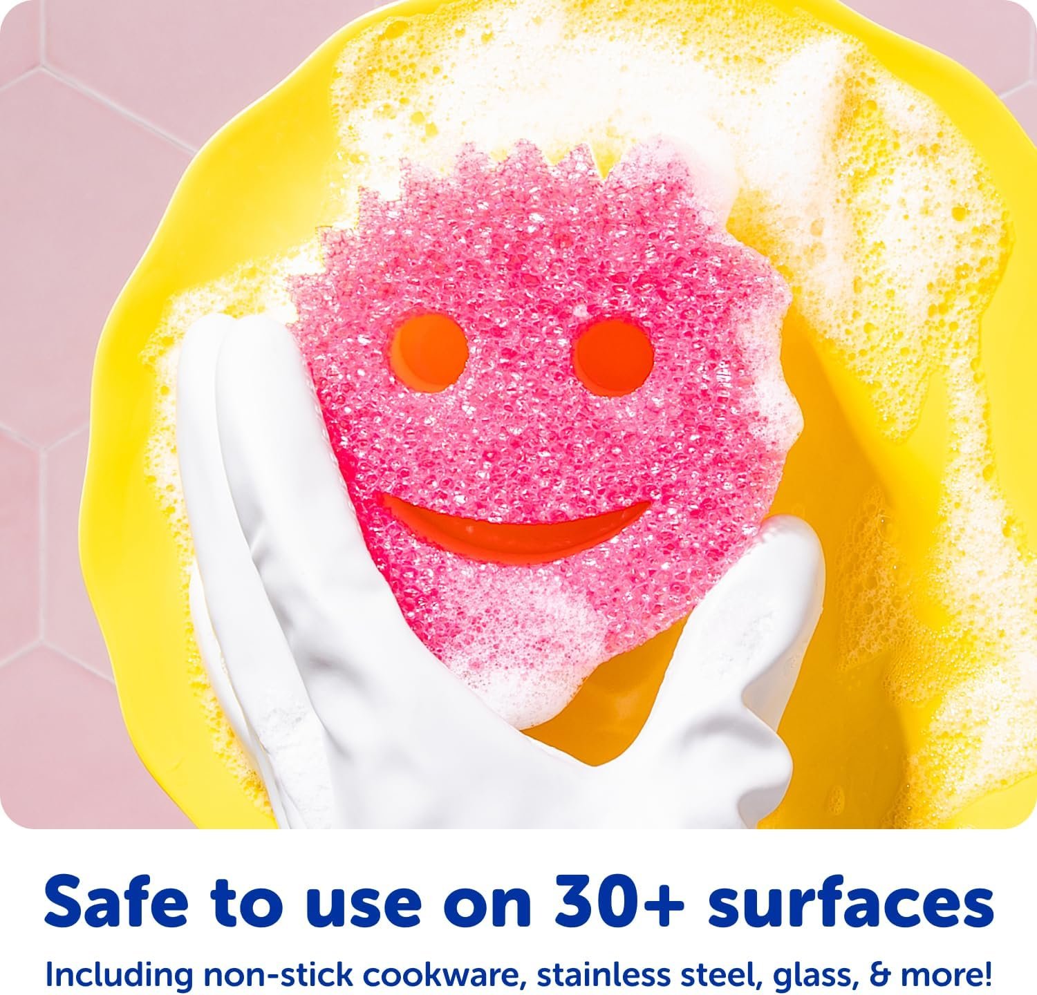 A colorful set of Scrub Daddy sponges (orange, green, pink) in a clear Daddy Caddy by a kitchen sink, with one sponge being rinsed under running water.