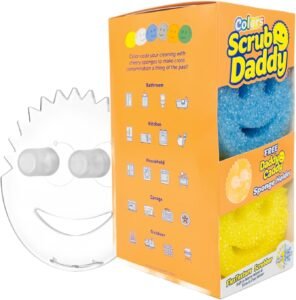 A colorful set of Scrub Daddy sponges (orange, green, and pink) in a clear Daddy Caddy by a kitchen sink, with one sponge being rinsed under running water.