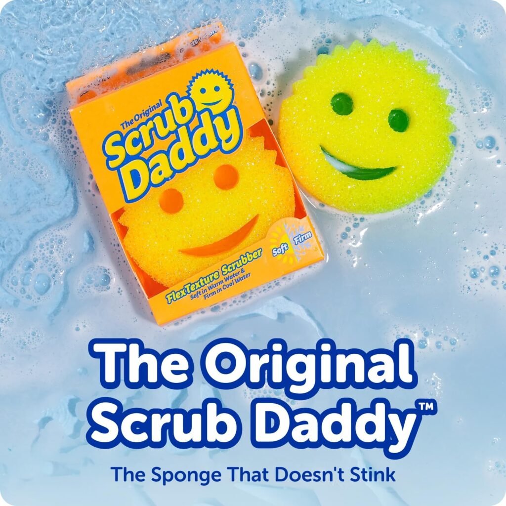 A colorful set of Scrub Daddy sponges (orange, green, and pink) in a clear Daddy Caddy by a kitchen sink, with one sponge being rinsed under running water.